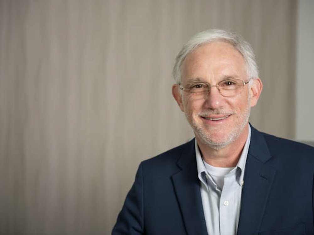 A head shot of a man with white hair and glasses in a dark suit looks at the camera