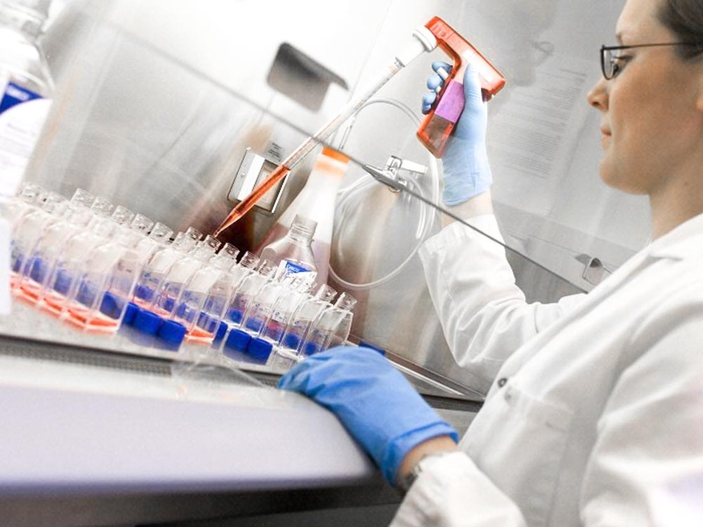 A person at a bench wearing a lab coat and gloves using a micro pipette