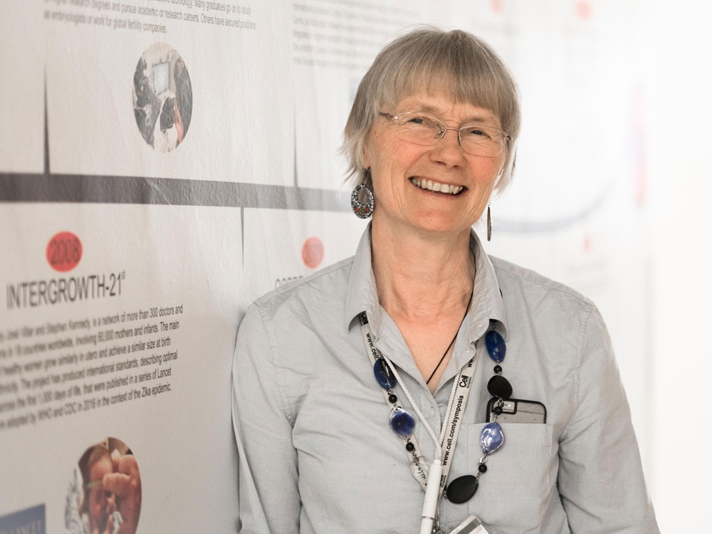 Jo Poulton - wearing a grey shirt and a chunky blue glass necklace, standing in front of a research information board