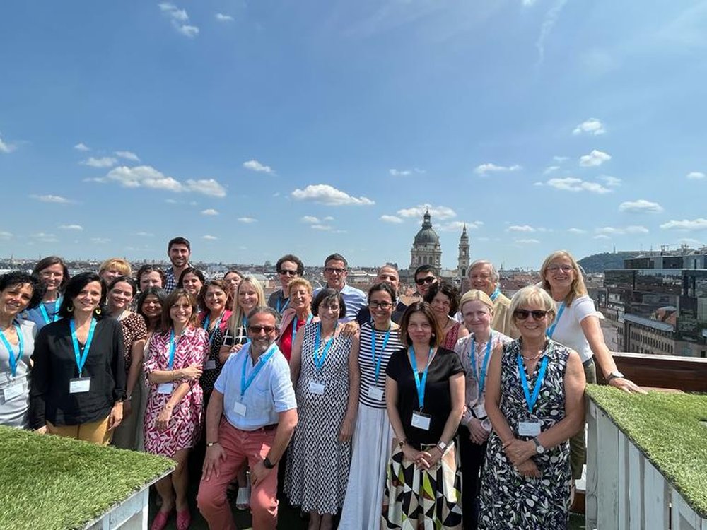 A group of people standing on the top of a building smiling, with a city and blue sky behind