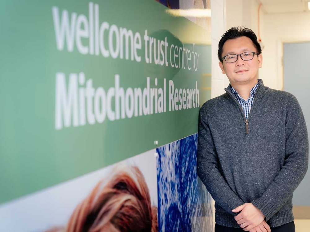 A man in glasses stands in front of a sign that says Welllcome Trust and Mitochondrial Disease