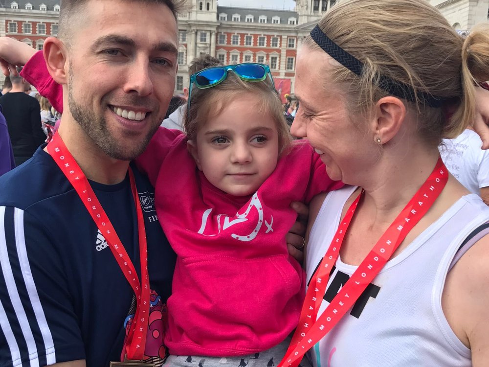 A man and a women hold between them a little girl in a pink Lily Foundation Jumper. They both are wearing medals