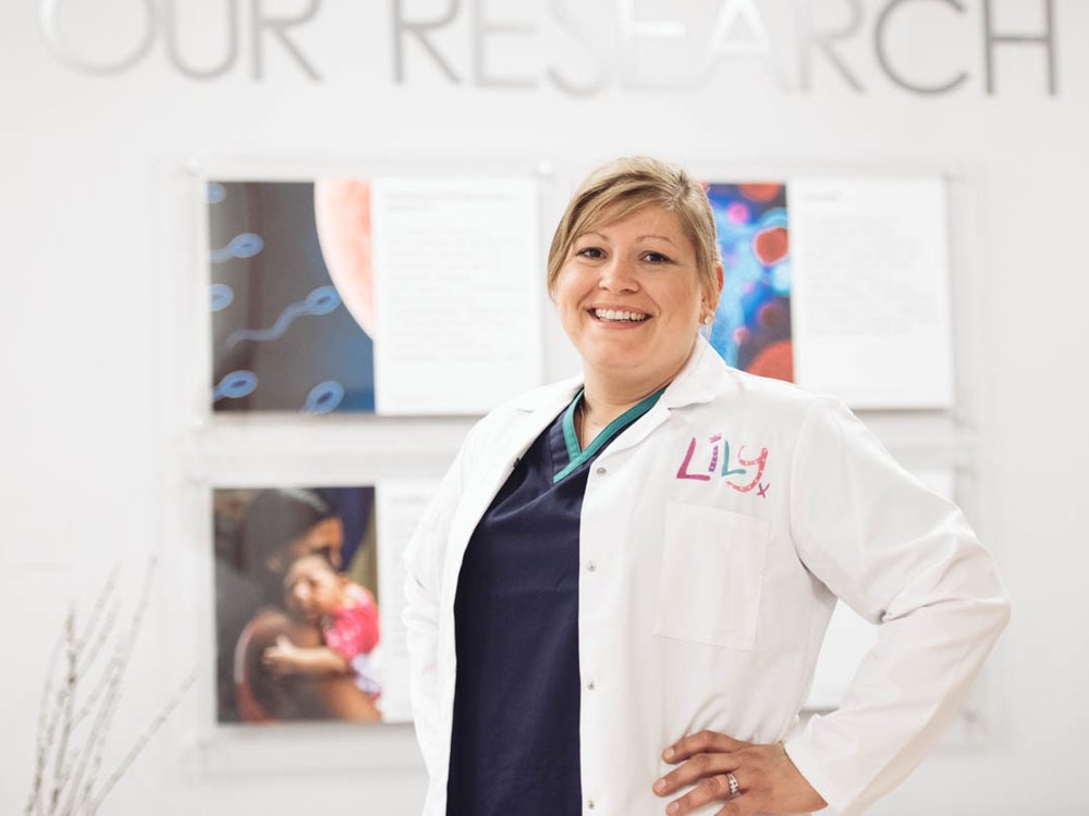 A women wearing blue scrubs and white Lily foundation lab coat stands with her hands on her hips smiling at the camera