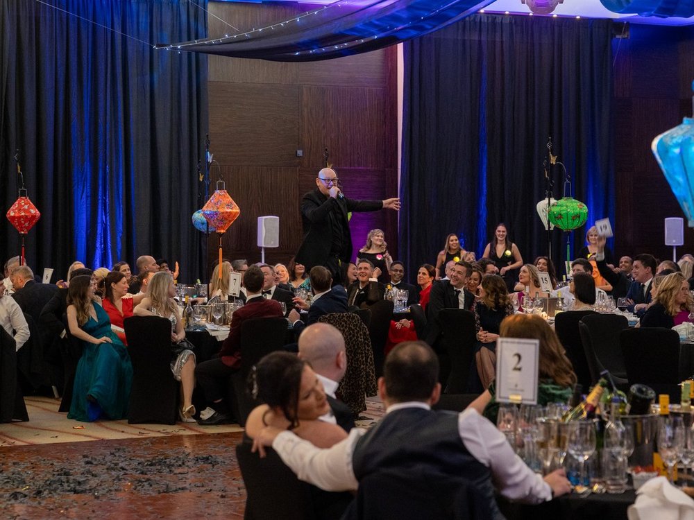 A man addresses a room full of people sat around tables