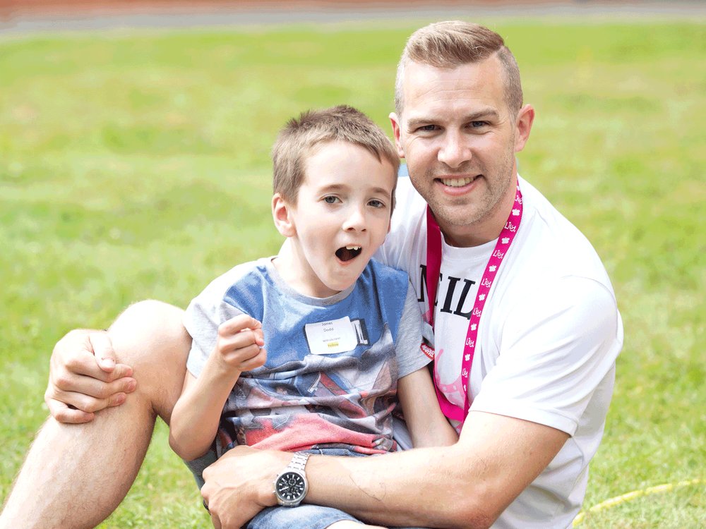 A man sits on the grass smiling at the camera with a small boy sat on his lap