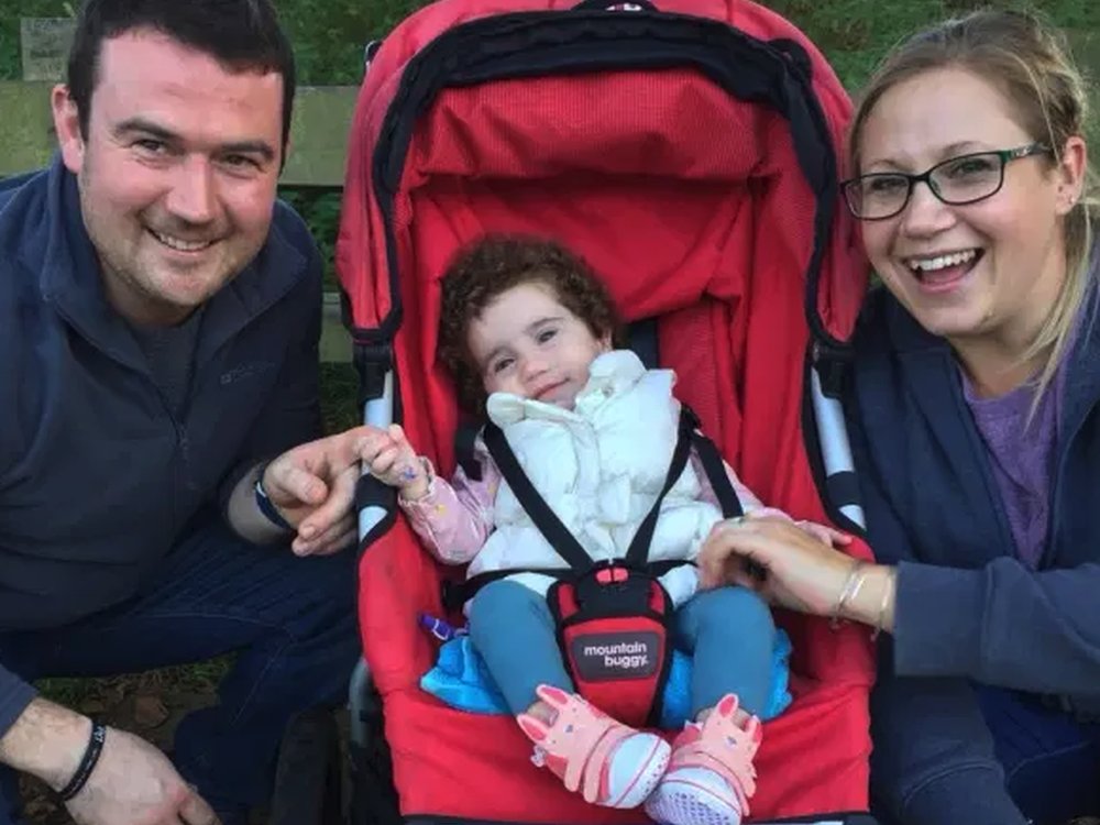 A man and a women crouching either side of a red pram smiling at the camera. In the pram is a little girl