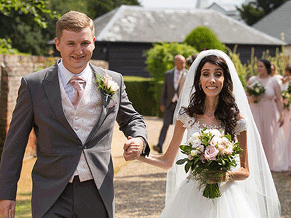 a man in a grey suit walks next to a women in a white wedding dress and veil holding flowers. Both are smiling