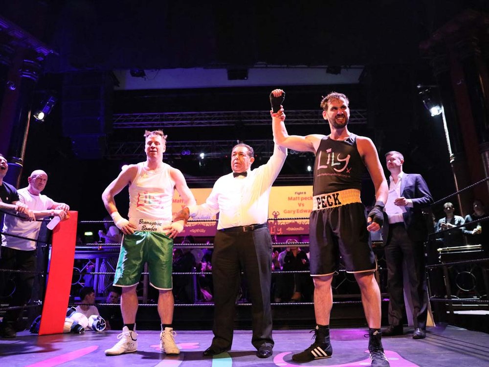 Two men in Lily Foundation vests and boxing shorts and gloves stand in a boxing ring. Between them is a referee .