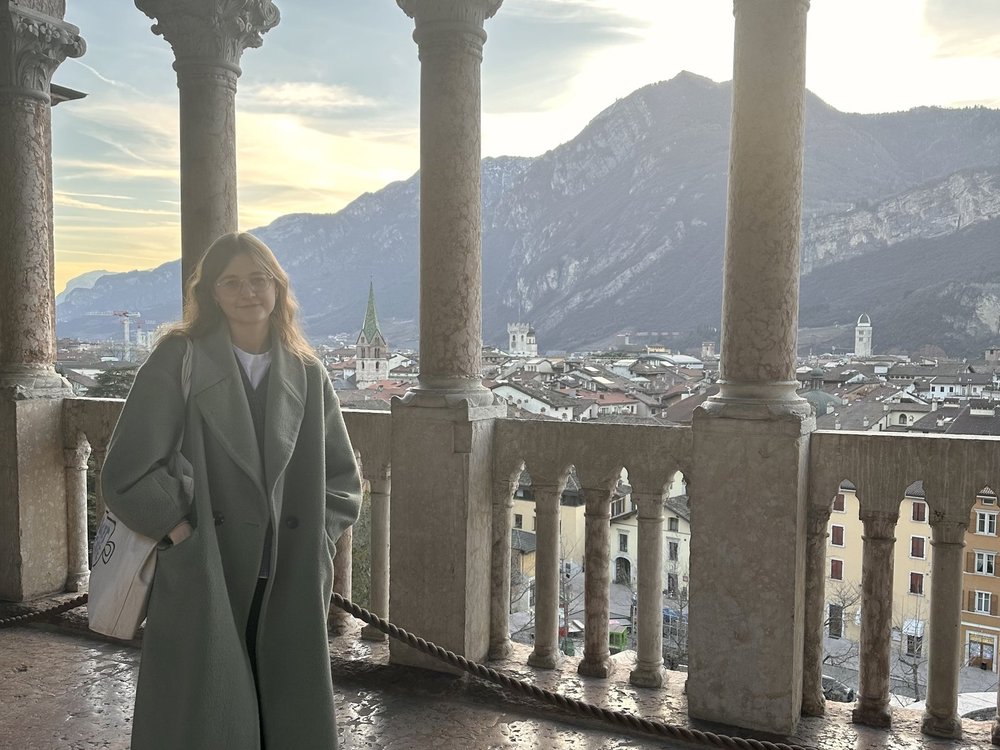 A lady standing on a balcony with colonnades, with a town and mountain scenery in the background