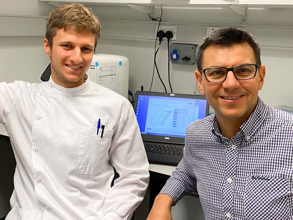 Two men stand In front of a laptop screen. One is in a lab coat the other is in a shirt