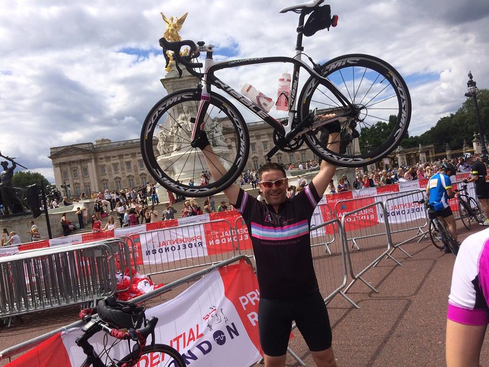 A man in a Lily cycling outfit holding a racing bike above his head