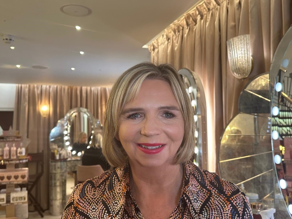 A lady with bobbed blonde hair smiling in a room with mirrors behind