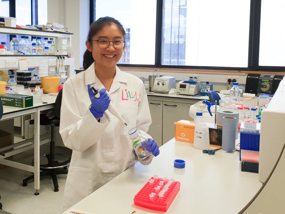 A women with dark hair and glasses wearing a Lily Foundation Lab coat smiles to the camera using a micro pipette