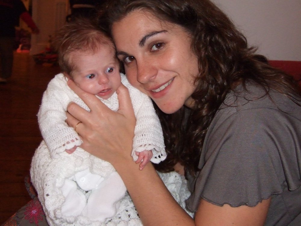 A lady with long dark hair cuddling a very young baby wrapped in a white knitted dress