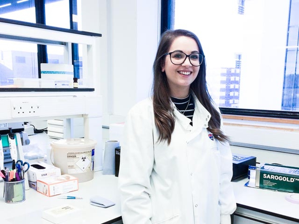 A woman with long dark hair in a lab coat stands in a laboratory