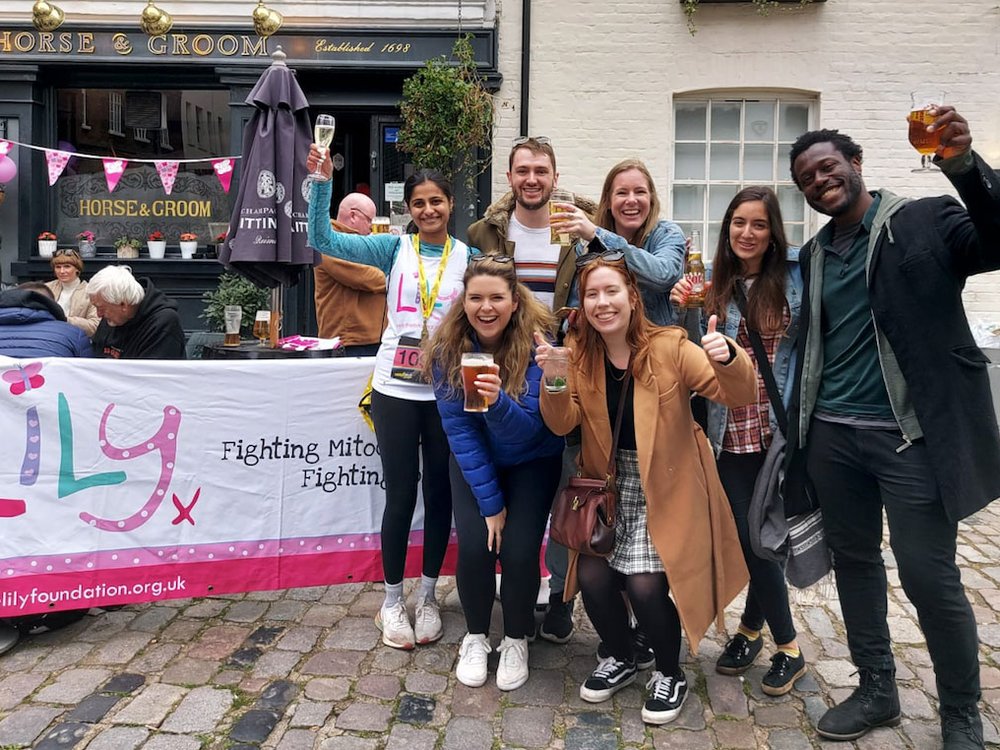 7 young adults stand outside a pub raising glasses. One is wearing a Lily running top and wearing a medal round her neck