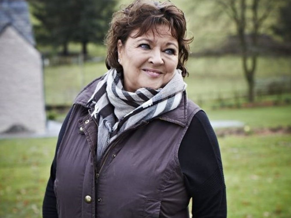 A lady with short dark hair standing in a field wearing a gilet and scarf, looking askance