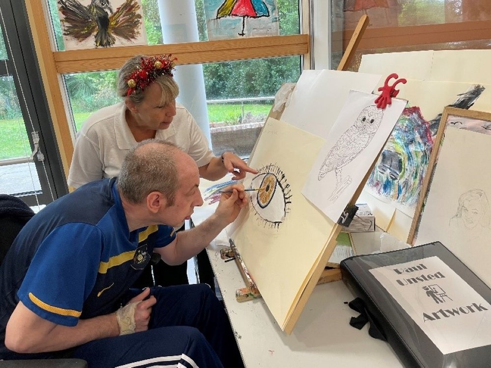 A man leans forward to paint on an easel while a woman stands next to him