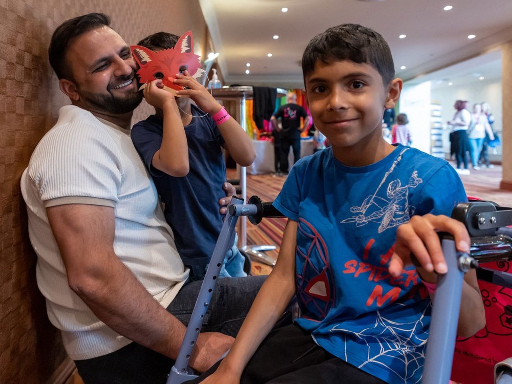 A man is sat on the floor smiling looking at a boy who is using a walker, on his lap is another child wearing a fox mask