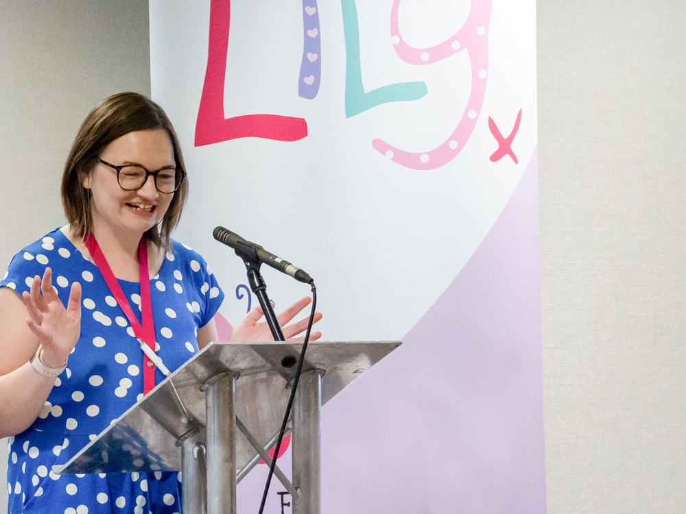 A woman stands in front of a lectern with a microphone attatched to it. Behind her is a sign with the Lily Foundation logo on