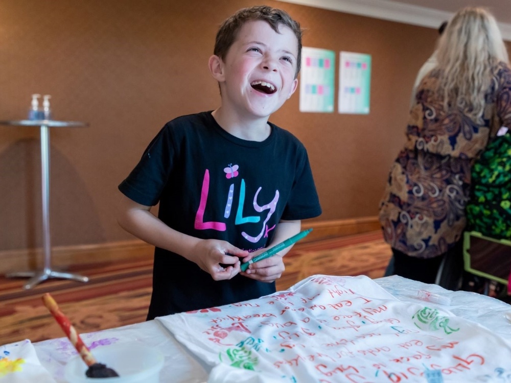 A little boy in a Lily Foundation t-shirt holds a pen. He is smiling and having fun