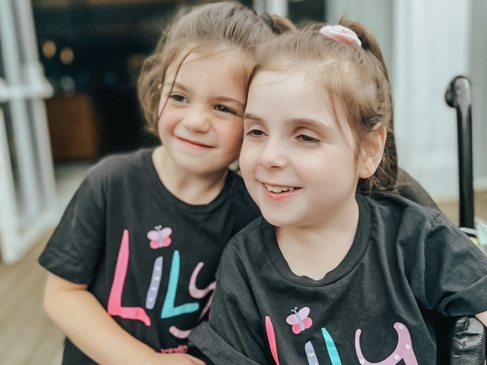 A little girl with mitochondrial disease in a wheel chair wearing a Lily Foundation tshirt. Stood beside her is a younger little