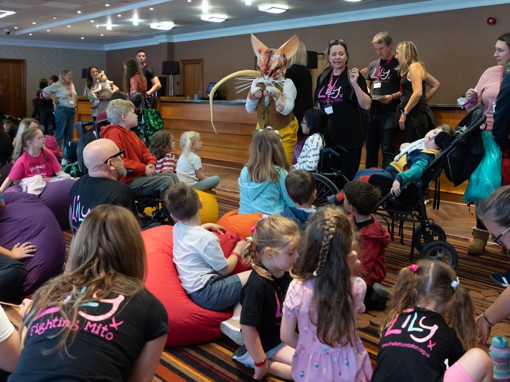 A group of children and Lily volunteers sitting watching someone dressed up in a mouse costume