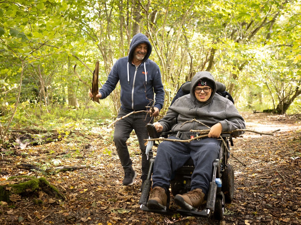 A mito patient in a wheelchair gather sticks in the forest, accompanied by a man