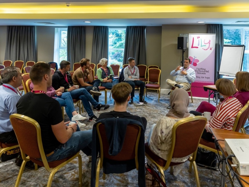 A group of adults sit on chairs in a circle listening to a man at the front speaking