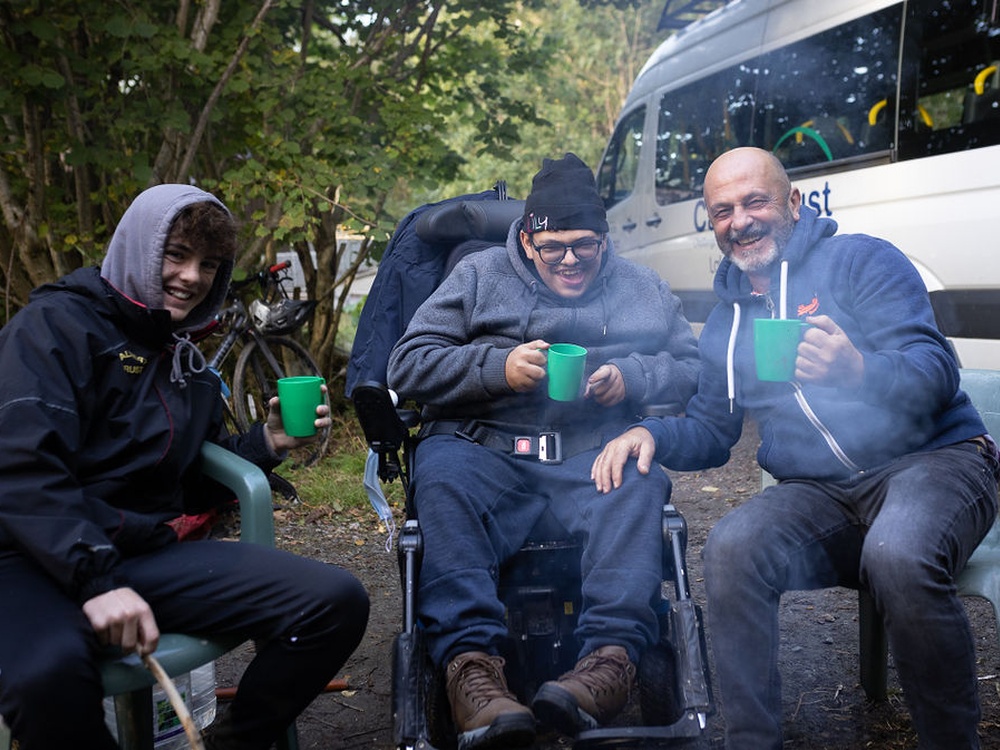 Three people sitting outside with cups in their hands, toasting to the camera